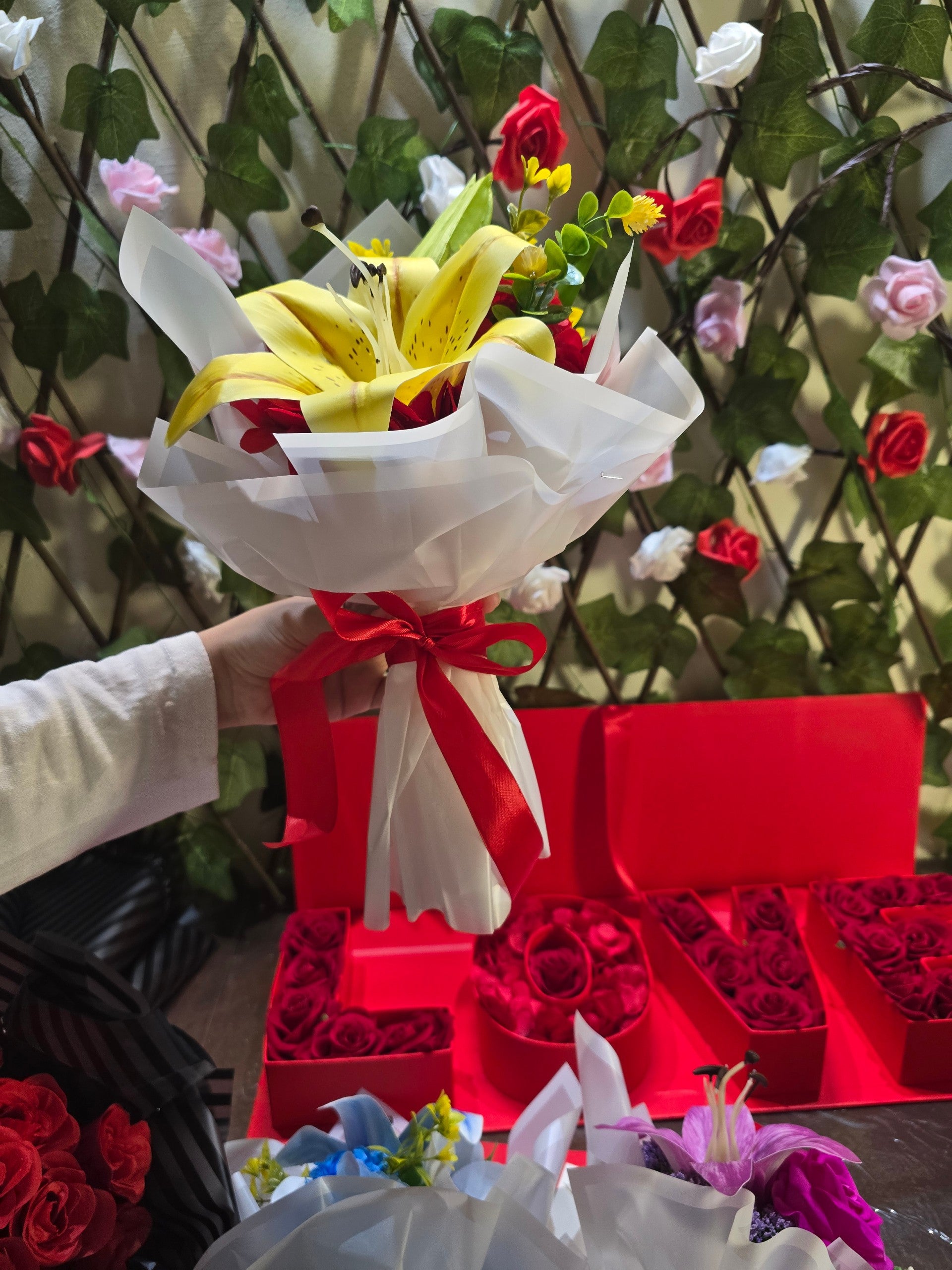 BioCode artificial yellow lily bouquet wrapped in white paper with a red ribbon, held in front of a rose-filled display