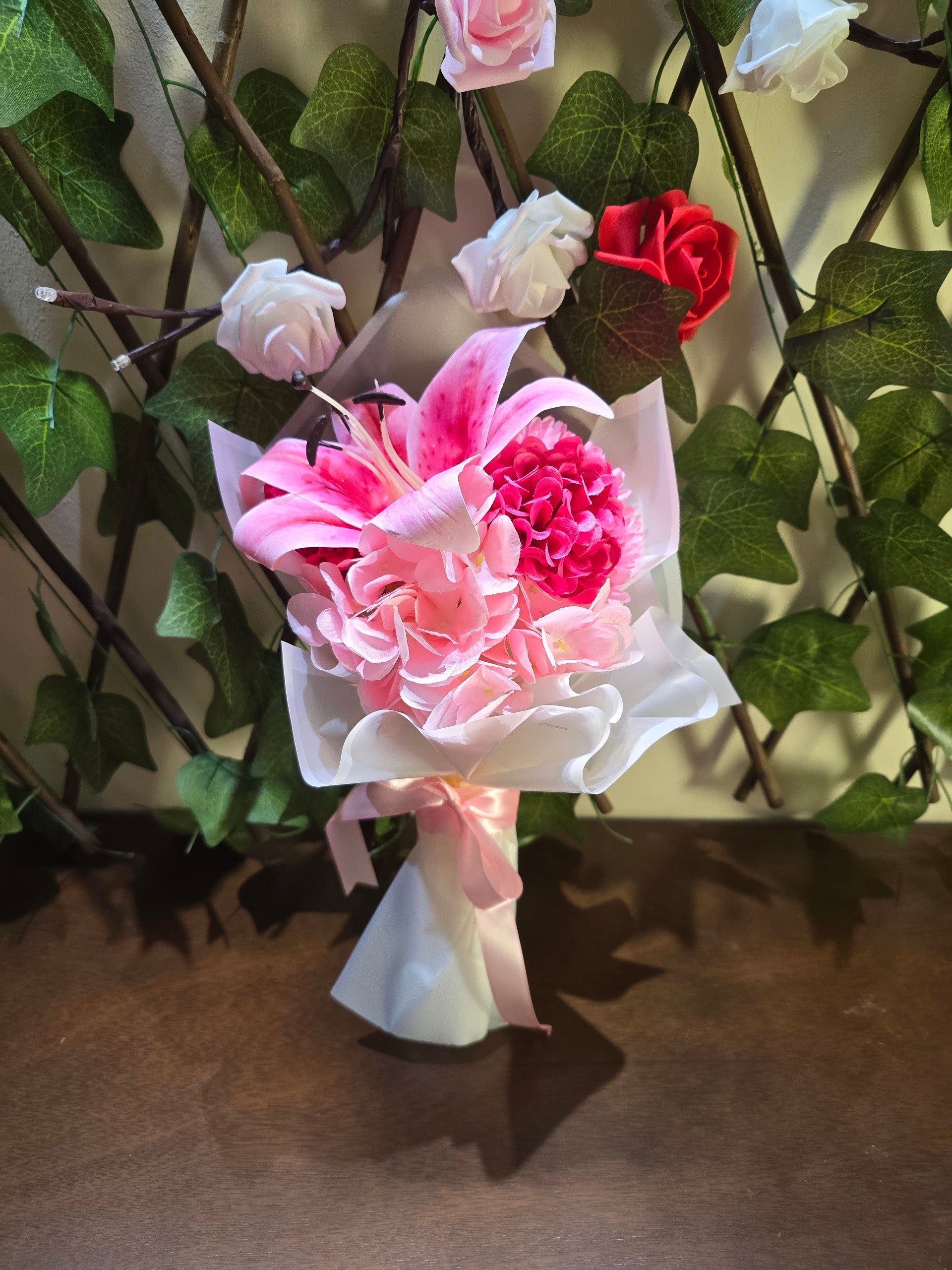 BioCode artificial flower bouquet featuring a pink lily and hydrangea blossoms wrapped in white paper with a pink ribbon