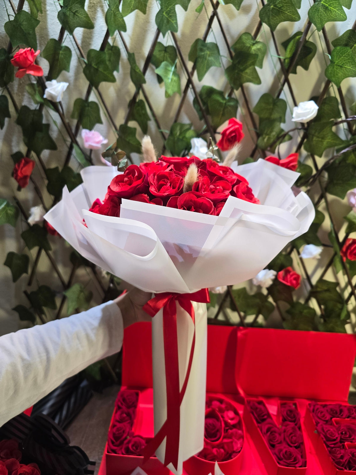 BioCode handcrafted artificial red rose bouquet wrapped in white paper with a red ribbon, held against a trellis.