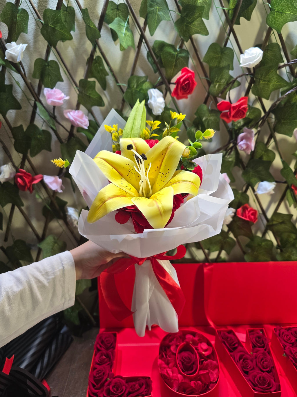 BioCode artificial yellow lily bouquet wrapped in white paper with a red ribbon, held against a floral trellis.