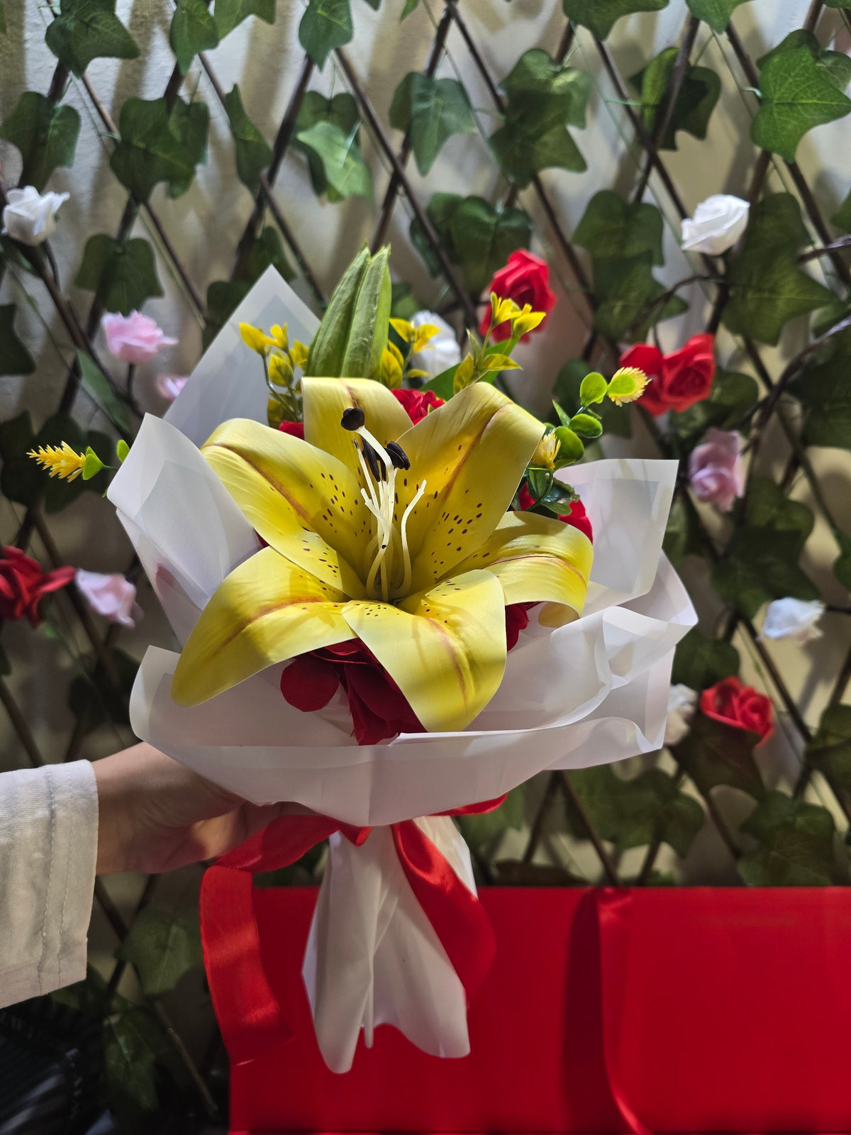 BioCode artificial yellow lily bouquet wrapped in white paper with a red ribbon, featuring red accents and greenery.
