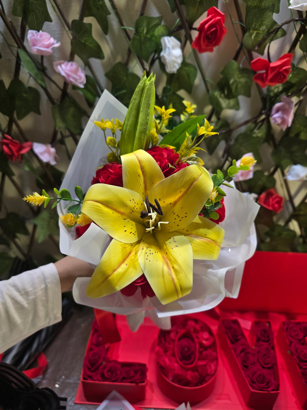 BioCode artificial yellow lily bouquet with red accents and greenery, wrapped in white paper with a red ribbon.