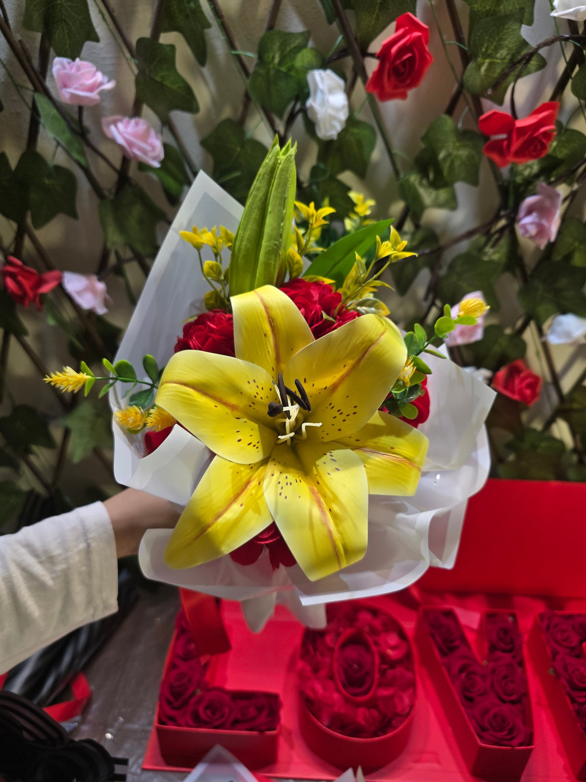 BioCode artificial yellow lily bouquet with red accents and greenery, wrapped in white paper with a red ribbon.