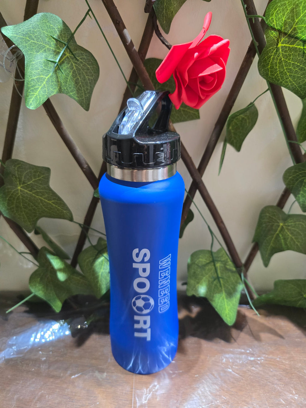 BioCode blue sports water bottle with black lid and WENEED SPORT branding, set against a backdrop of faux ivy and a red rose.