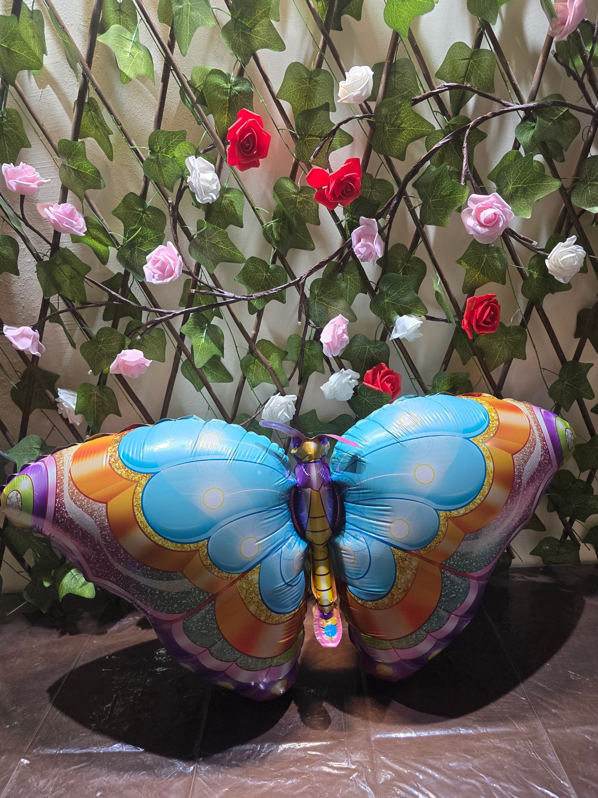BioCode large colourful butterfly balloon with shimmering patterns displayed against a decorative rose trellis wall