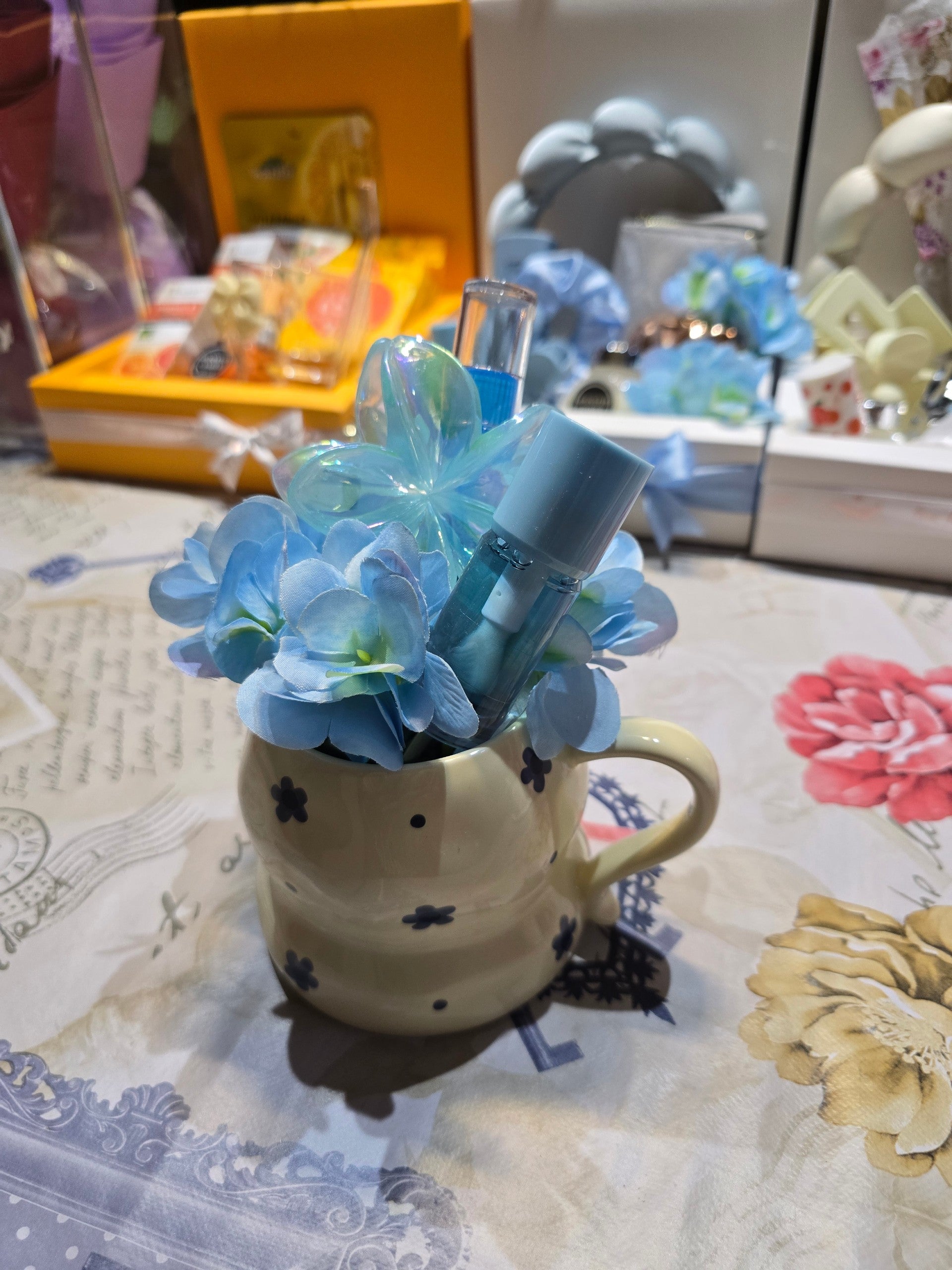 BioCode decorative cream ceramic mug filled with blue silk hydrangeas, an iridescent flower clip, and beauty items.