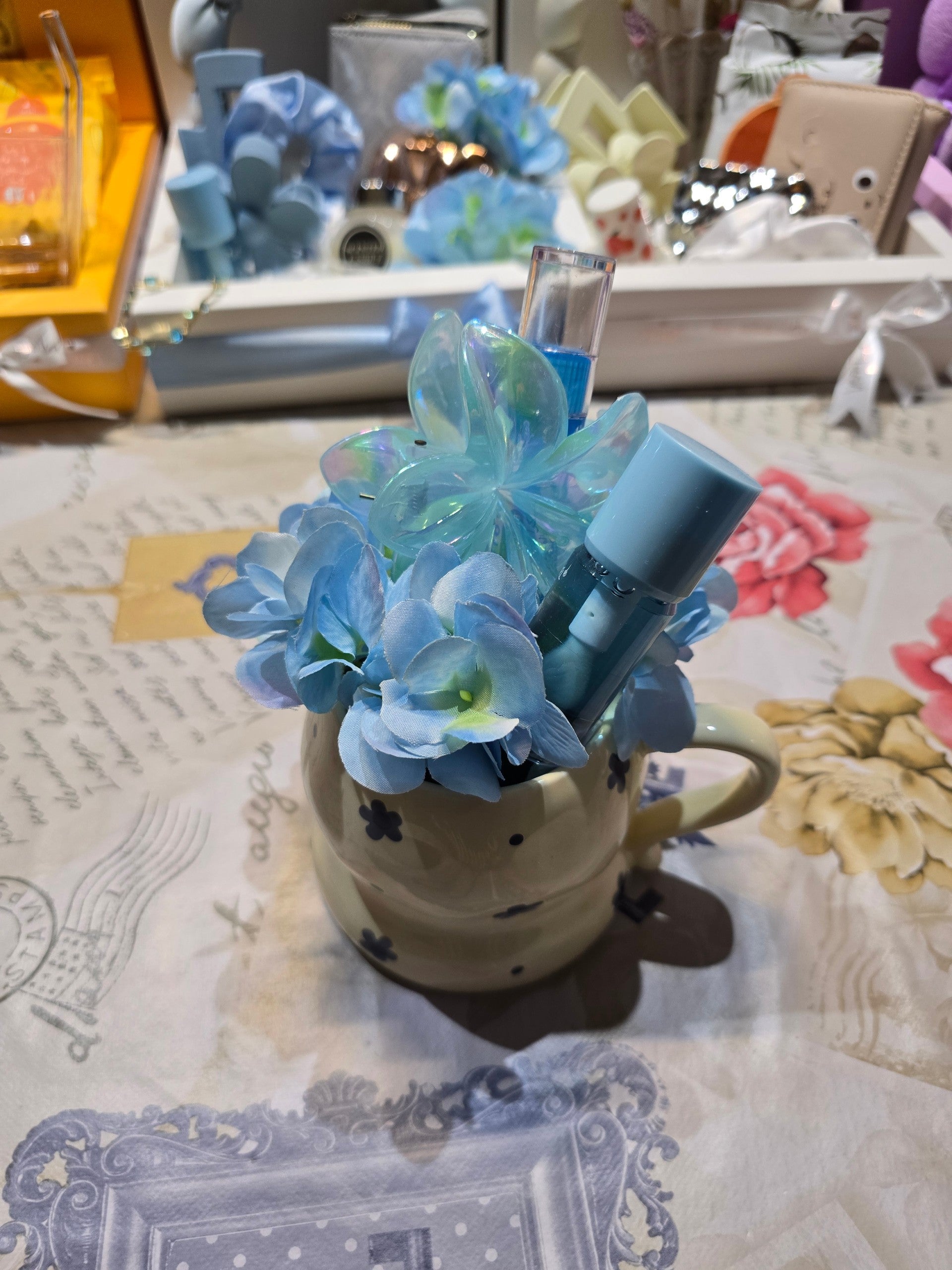 BioCode decorative cream ceramic mug filled with blue silk hydrangeas, an iridescent flower clip, and beauty items.