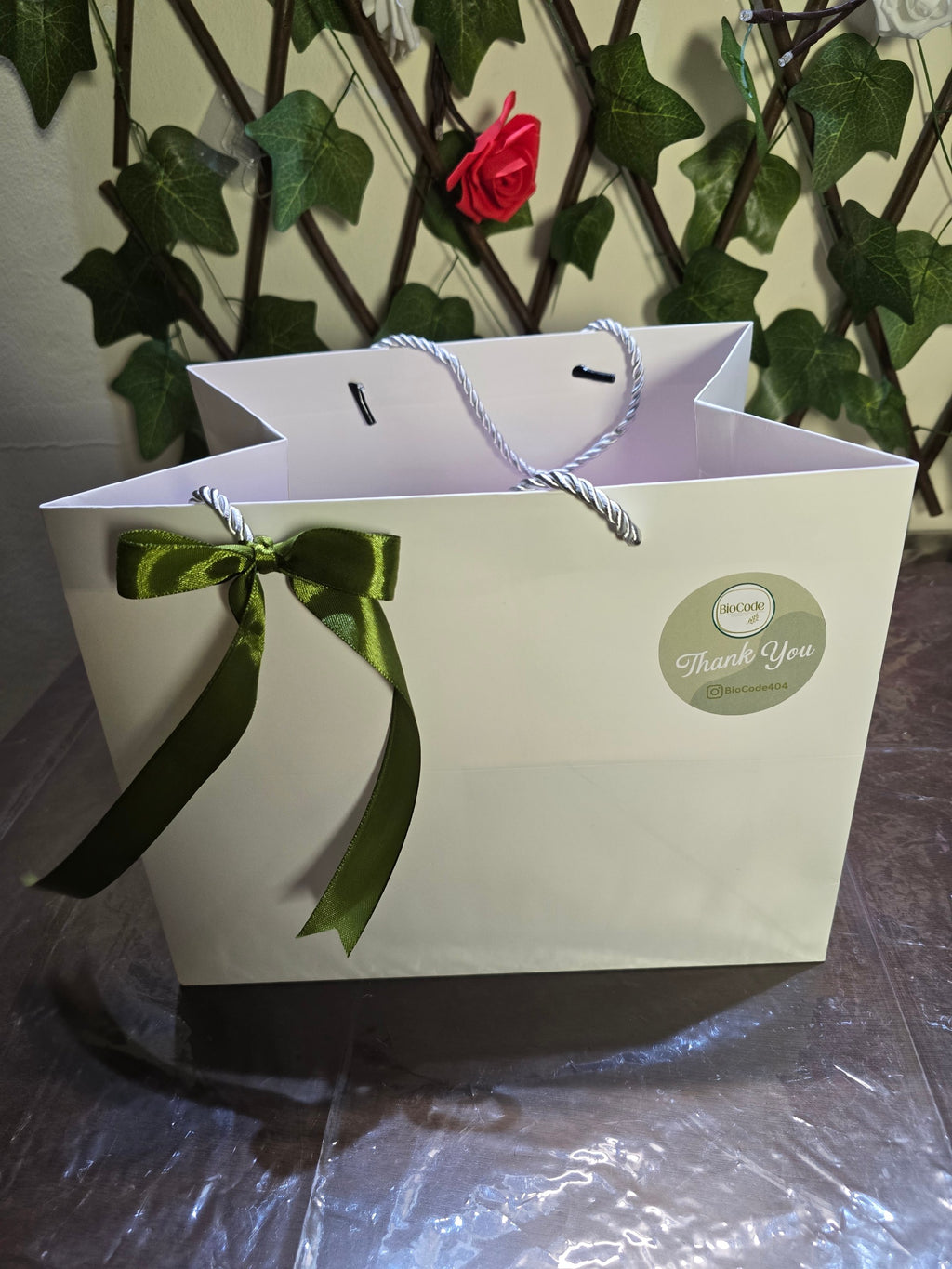 BioCode gift bag in white with a green satin ribbon bow and a Thank You sticker, set against a leafy wall background.