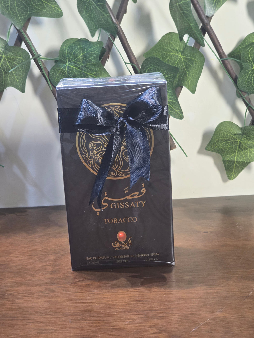 BioCode Gissaty Tobacco Eau de Parfum in a black gift box with a black ribbon, set against a backdrop of green leaves.