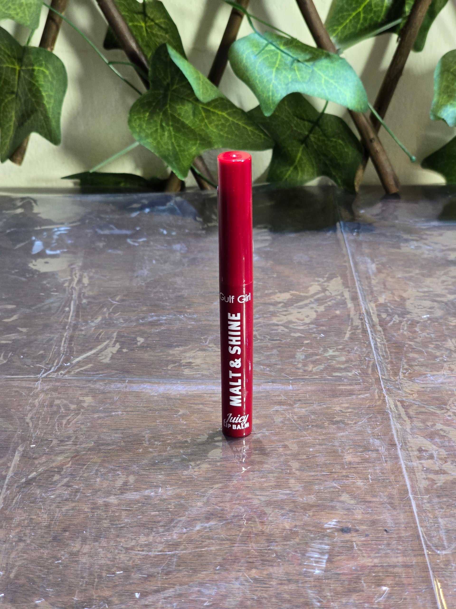 BioCode Gulf Girl Malt & Shine red lip balm tube standing upright on a reflective surface with green leaves behind.