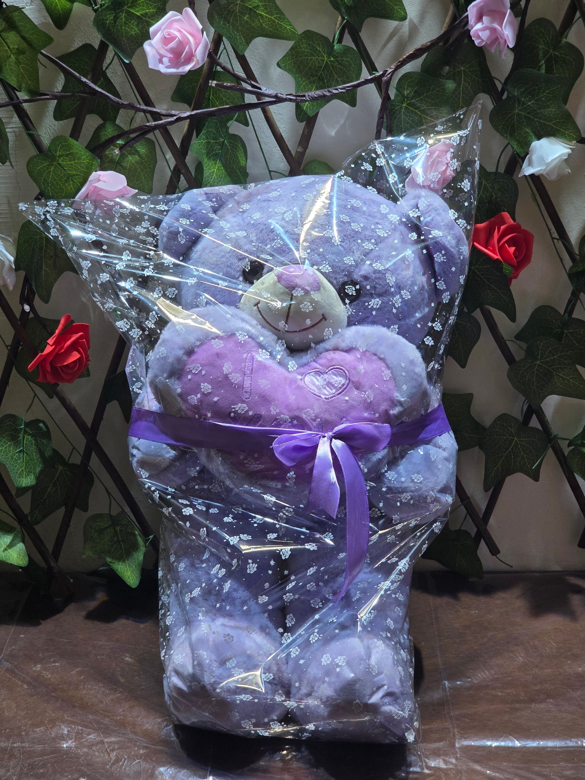 BioCode lavender plush teddy bear holding an I Love You heart, wrapped in clear floral cellophane with a purple bow.
