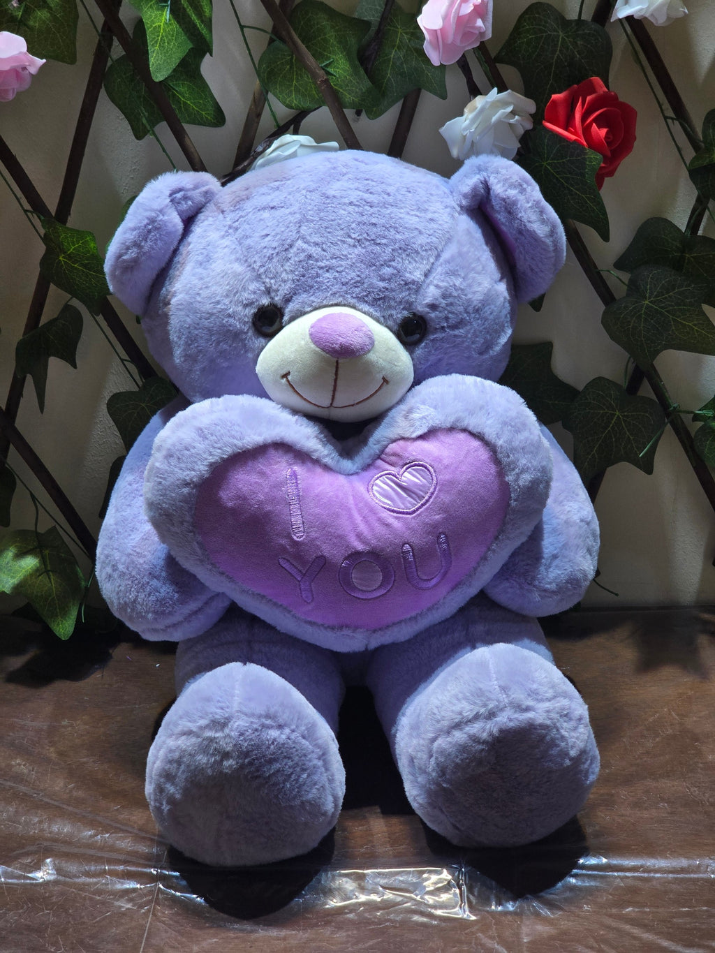 BioCode lavender plush teddy bear holding a soft heart pillow with I Love You embroidery against a floral backdrop