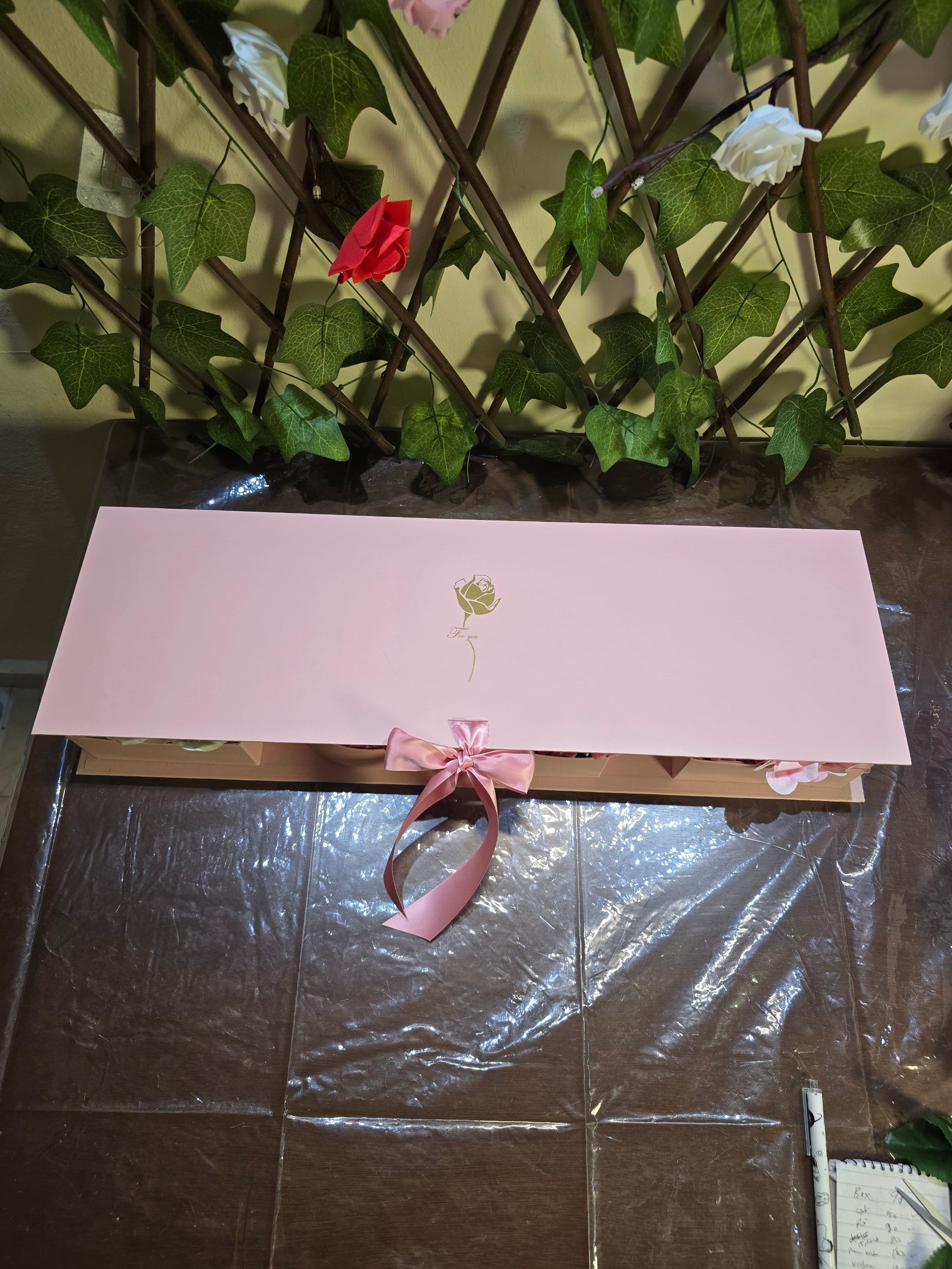 BioCode pink rectangular gift box with a gold rose emblem and satin ribbon, set against a decorative trellis backdrop.