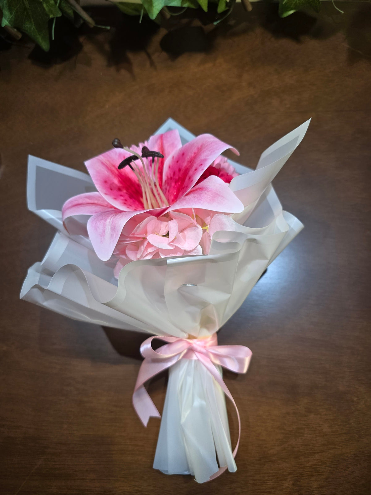 BioCode artificial pink lily and hydrangea bouquet wrapped in white paper with a pink satin ribbon on a wooden table