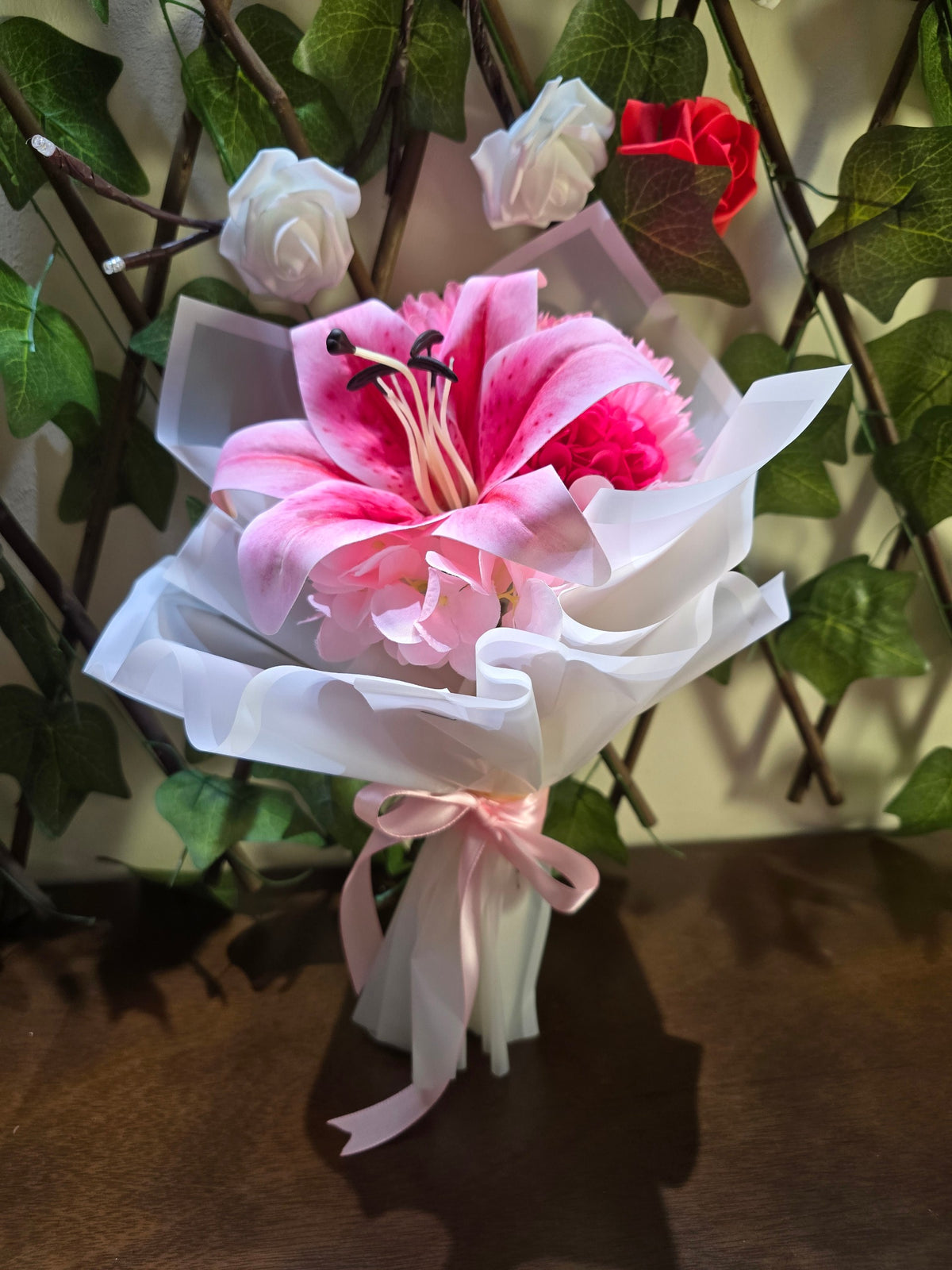 BioCode artificial pink lily bouquet wrapped in white paper with a pink satin ribbon, set against ivy trellis.