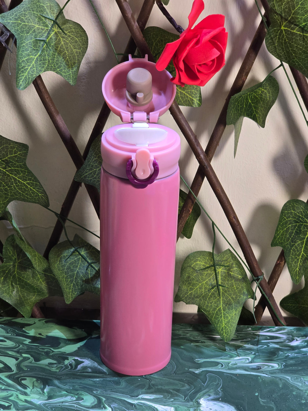 BioCode pink stainless steel vacuum insulated travel flask with a leak-proof flip-top lid, set against ivy foliage.
