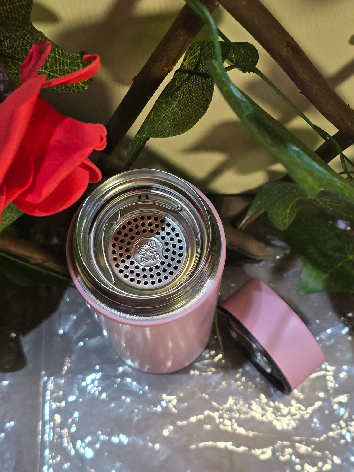 BioCode pastel pink stainless steel water bottle with internal tea infuser and lid, set against floral decor.
