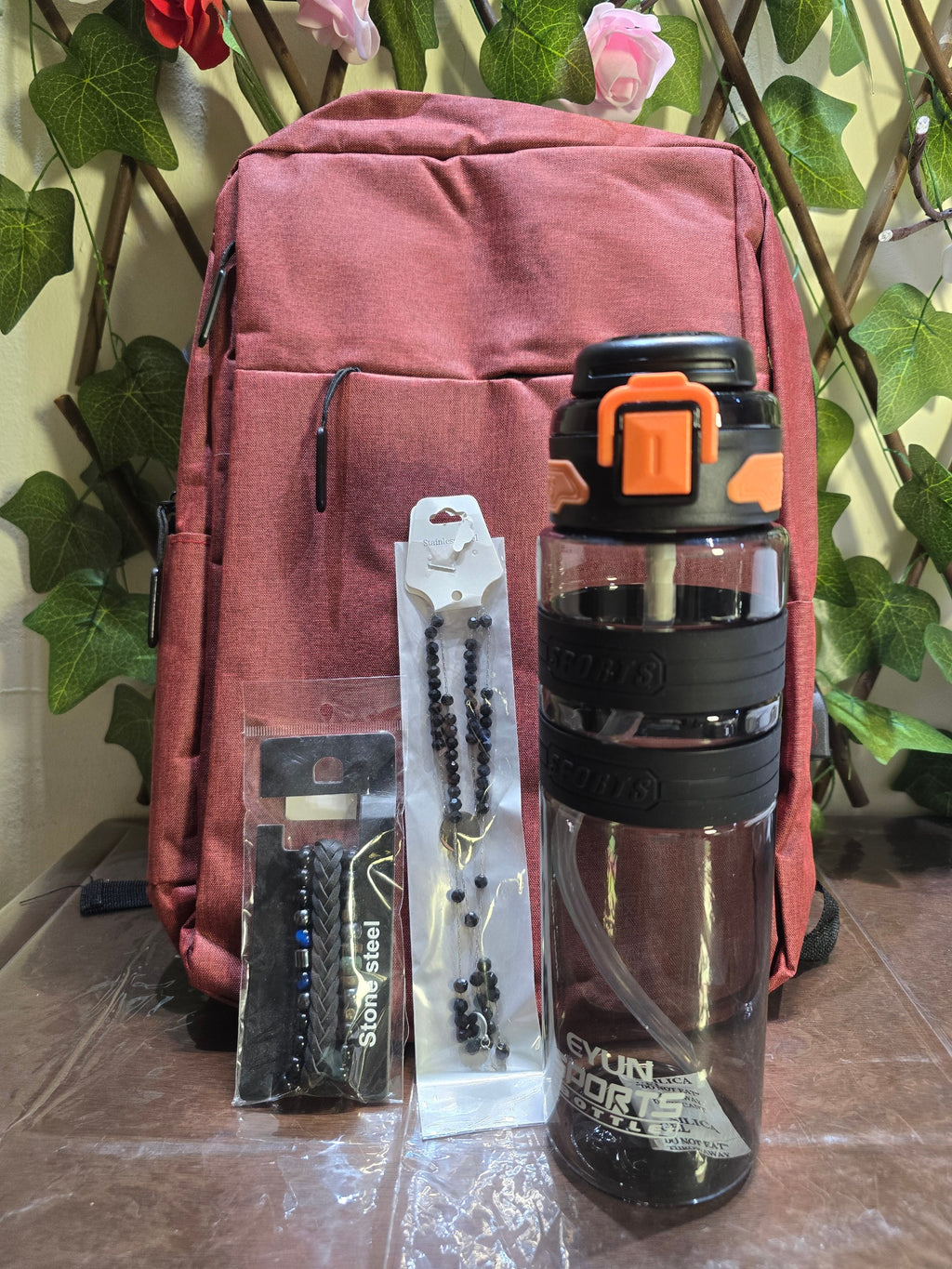 BioCode deep red laptop backpack with a sports water bottle and beaded bracelets in front of a green leaf backdrop.