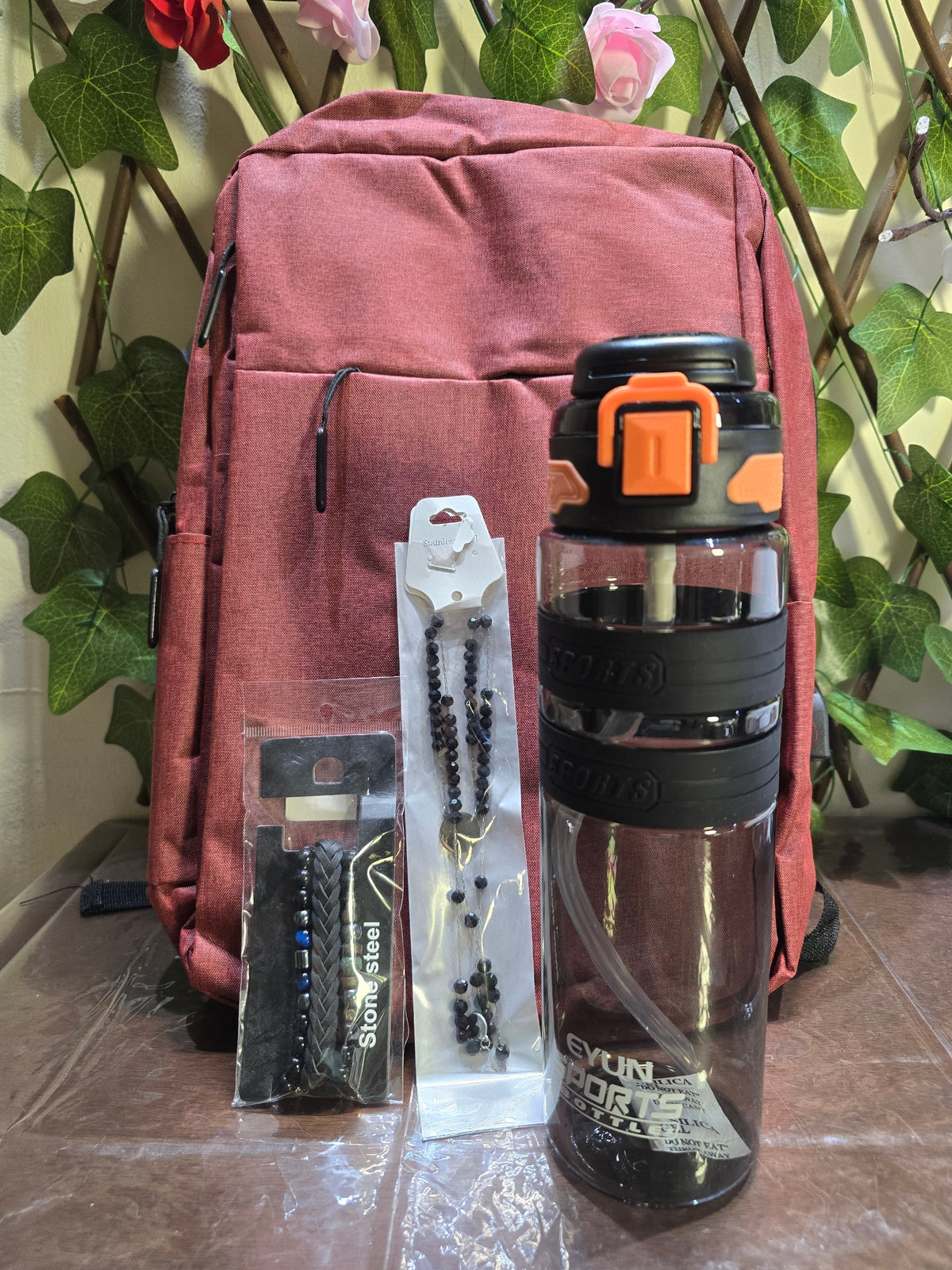 BioCode deep red laptop backpack with a sports water bottle and beaded bracelets in front of a green leaf backdrop.