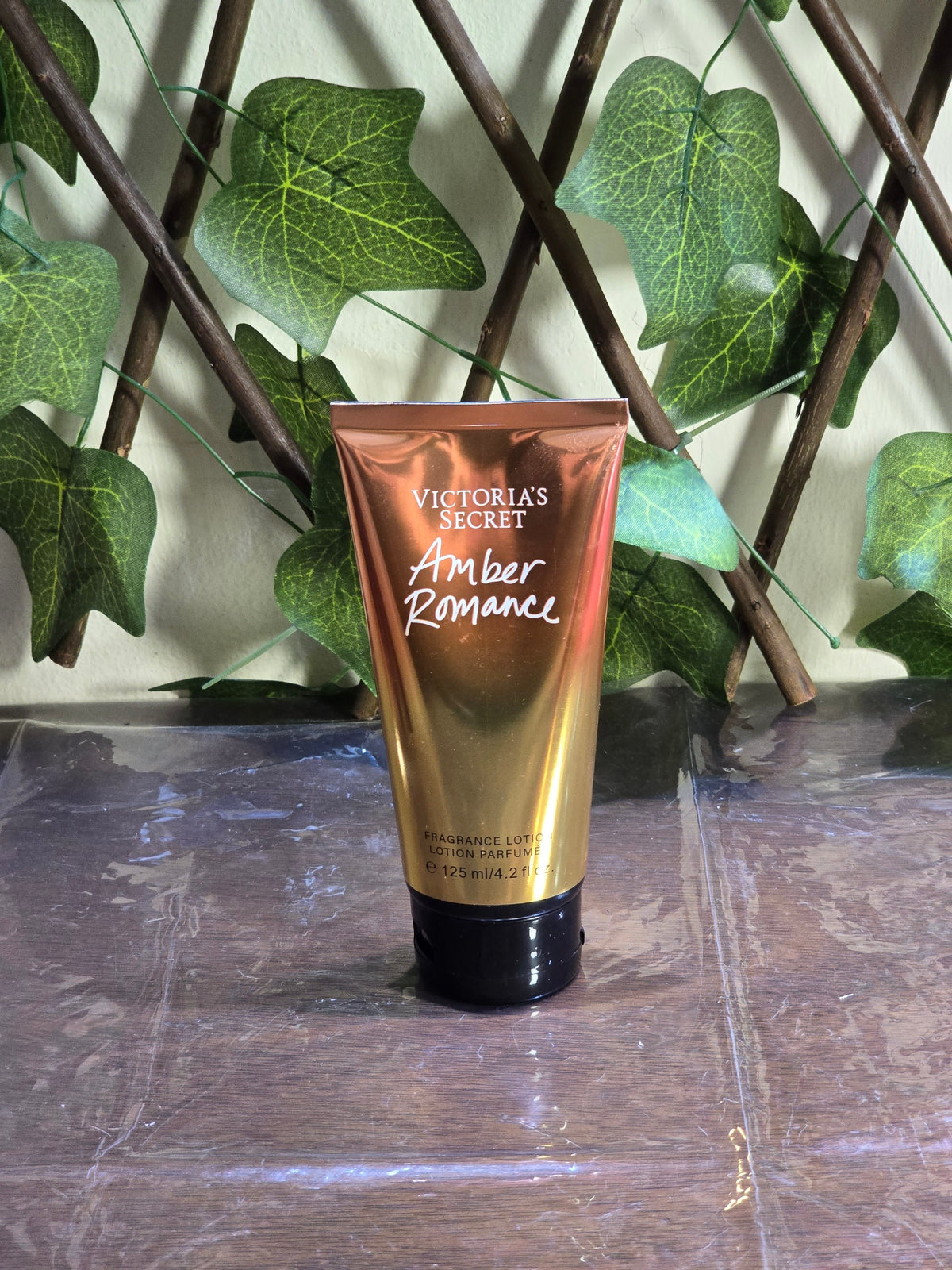 BioCode Victoria's Secret Amber Romance fragrance lotion in a metallic gold tube against a green ivy trellis background
