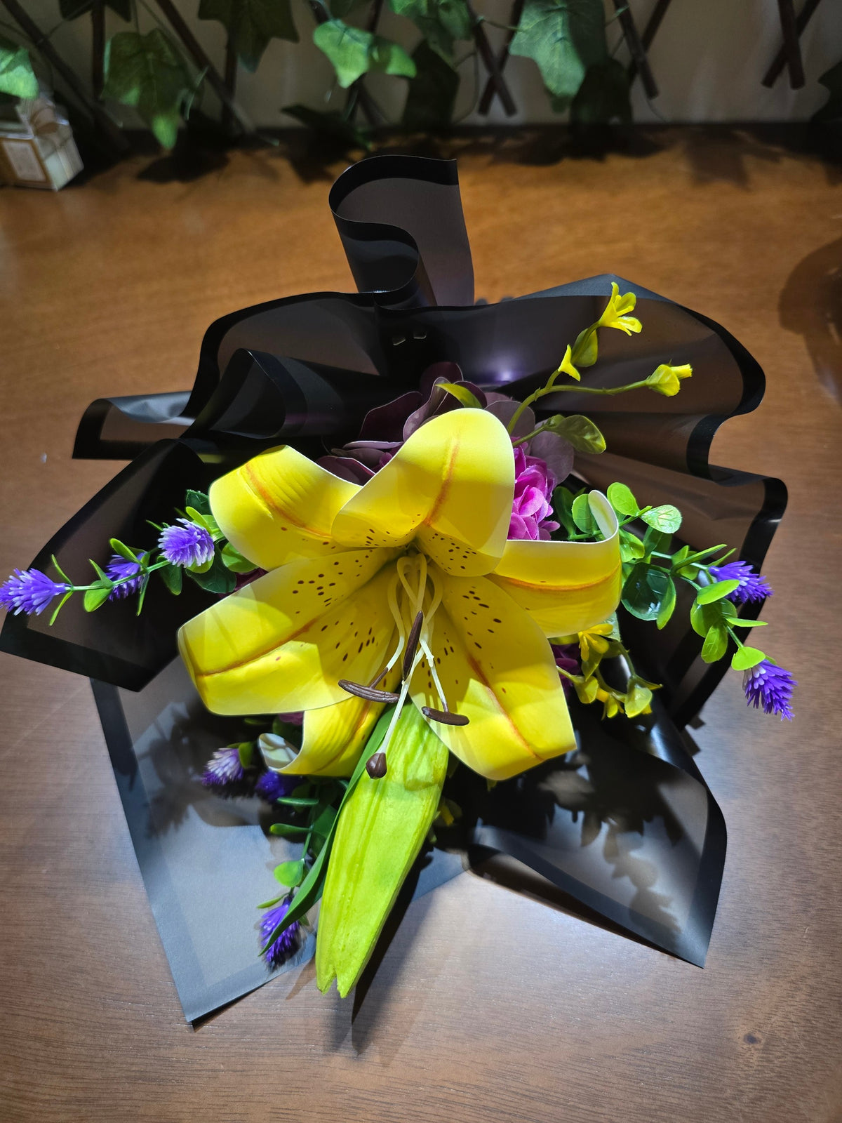 BioCode gift bouquet featuring a large yellow lily, purple accents, and green foliage wrapped in elegant black paper.