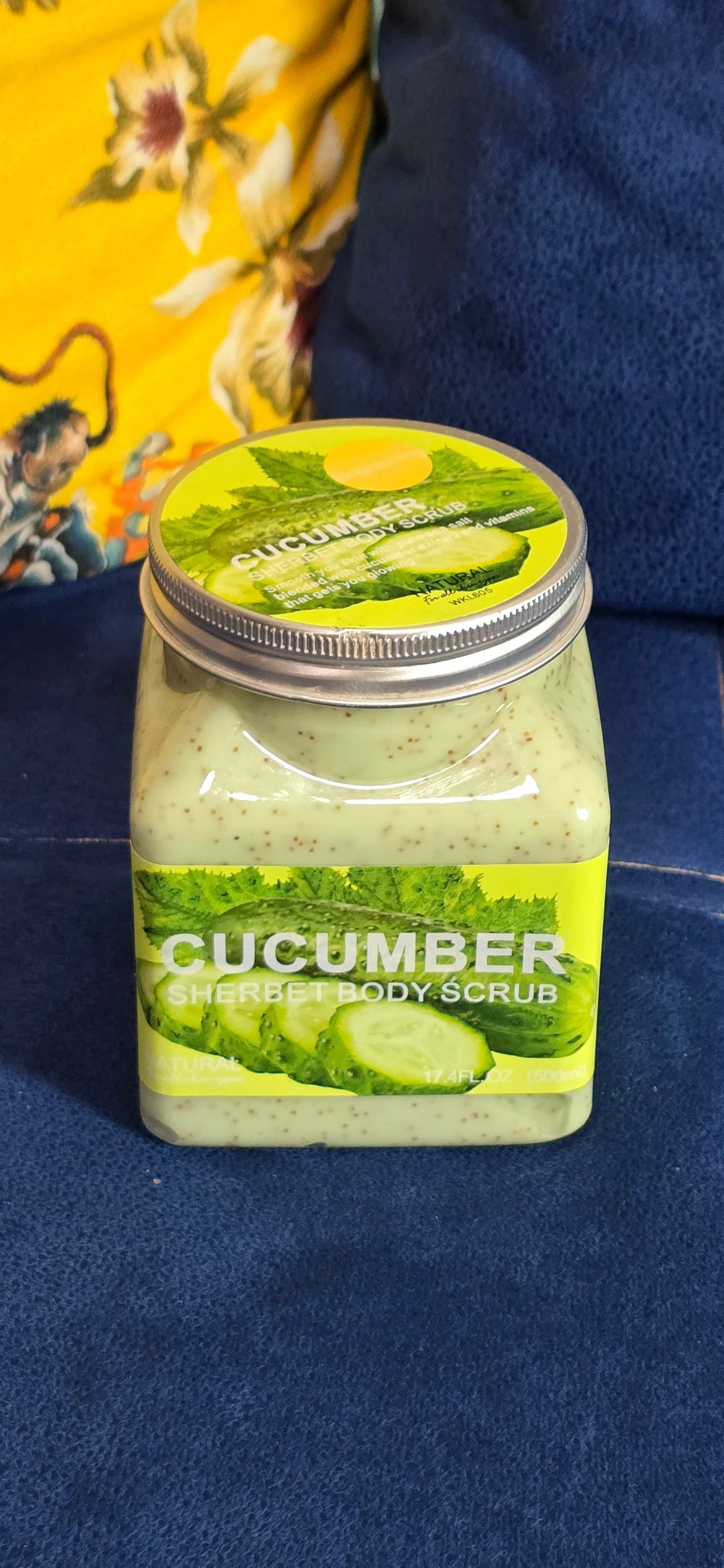 A jar of cucumber body scrub with a green and yellow label, resting on a dark blue textured surface.